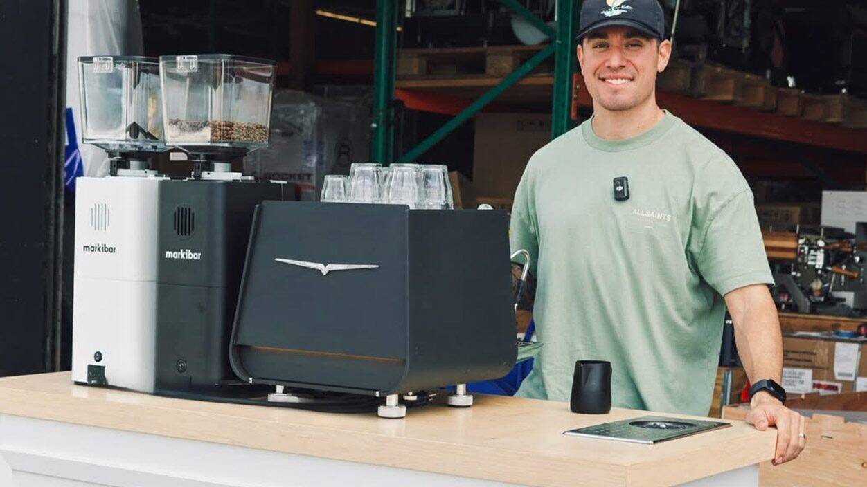 Coffee Machine Depot team member stands behind a demo counter with Markibar grinders and an espresso machine in a warehouse showroom.