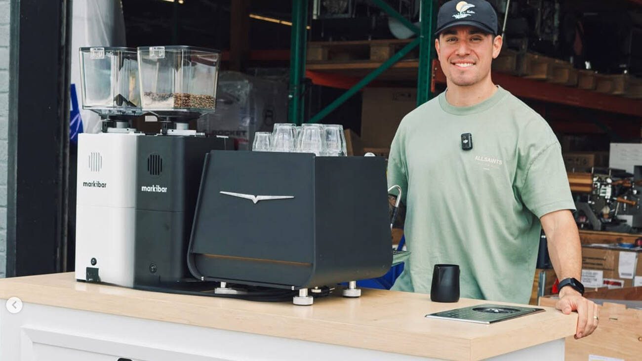 Coffee Machine Depot team member stands behind a demo counter with Markibar grinders and an espresso machine in a warehouse showroom.