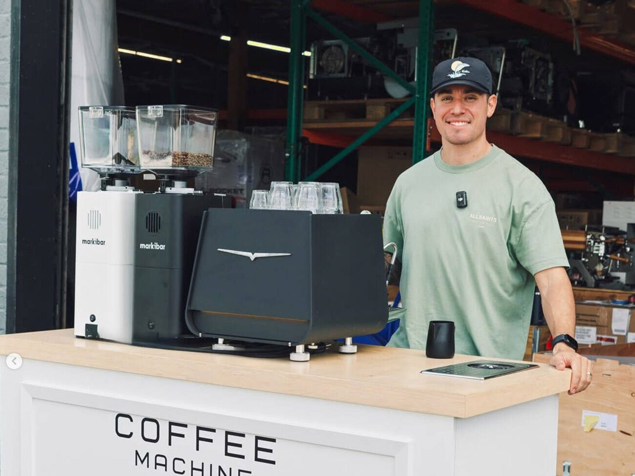 Coffee Machine Depot team member stands behind a demo counter with Markibar grinders and an espresso machine in a warehouse showroom.