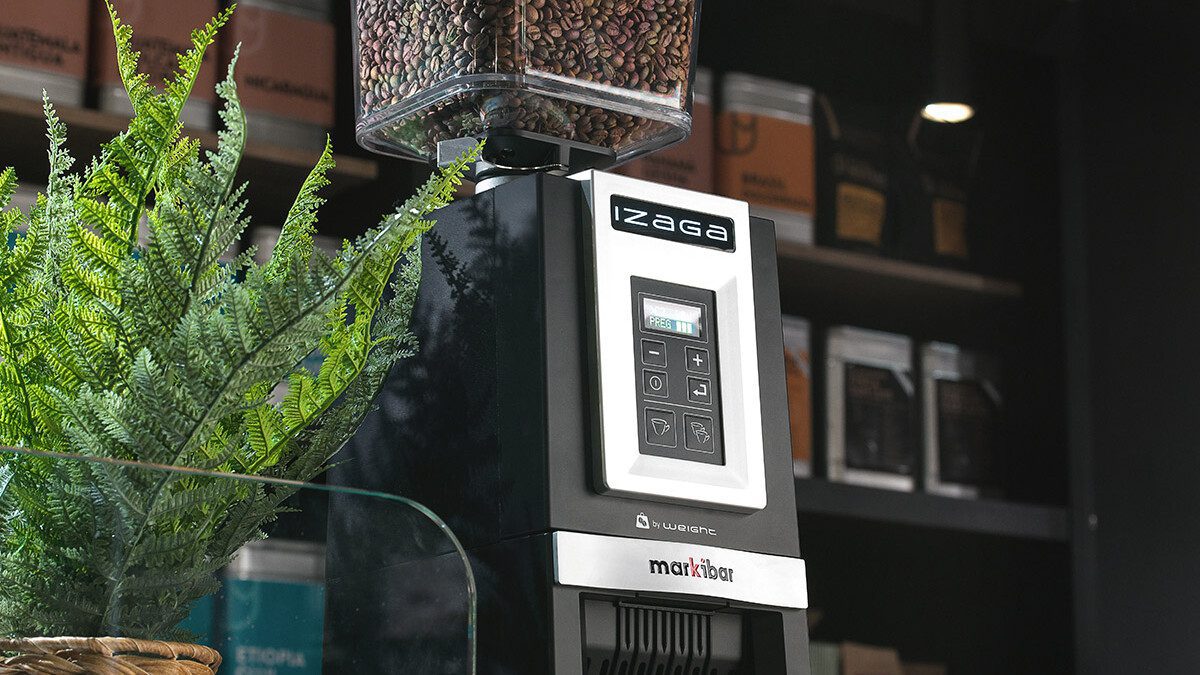 Markibar Izaga espresso grinder with a clear bean hopper on a café counter next to a potted plant, with coffee bags displayed in the background.