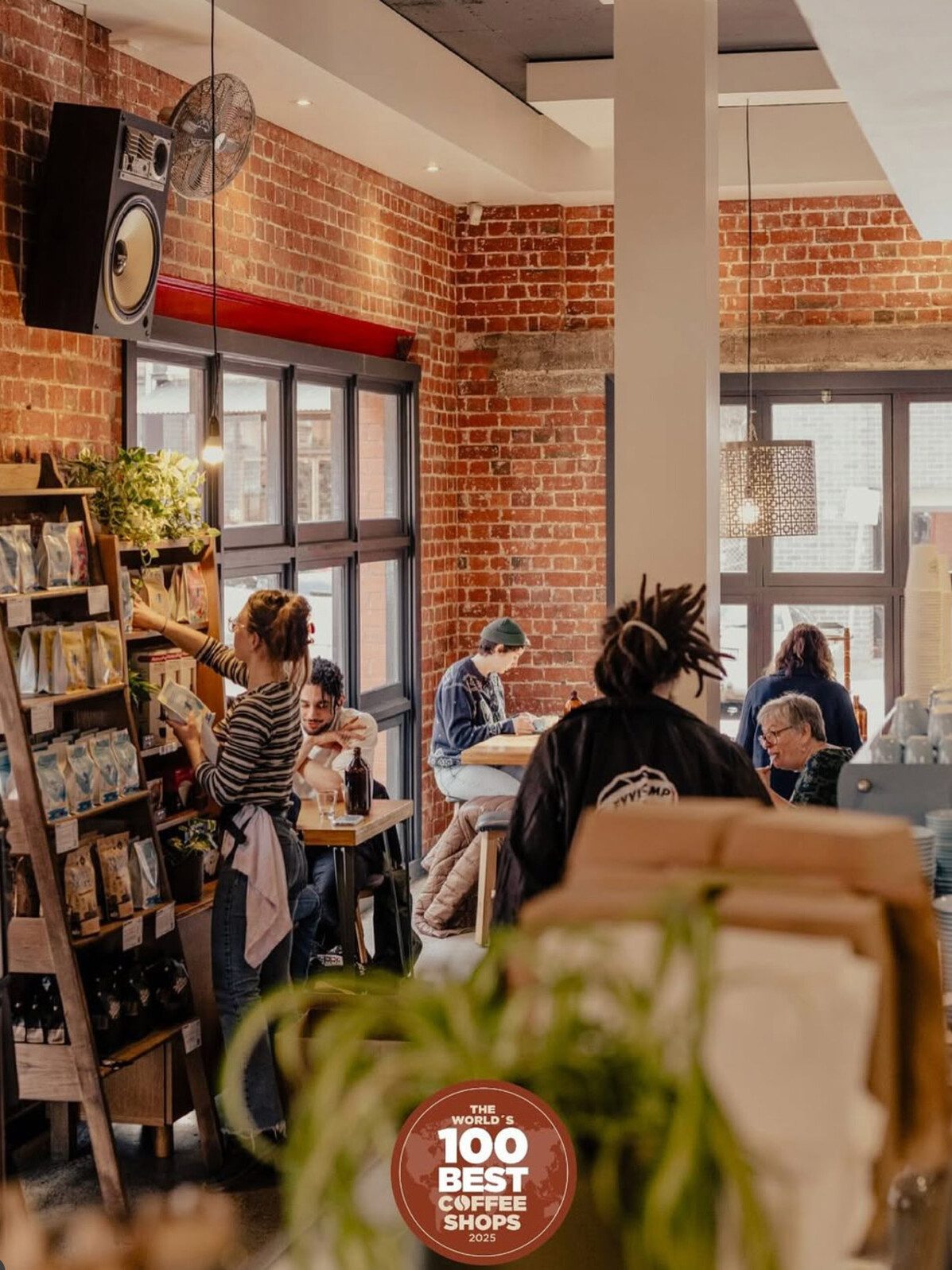 Cozy brick-walled coffee shop interior with customers seated at tables and a barista organizing coffee bags on a display shelf.