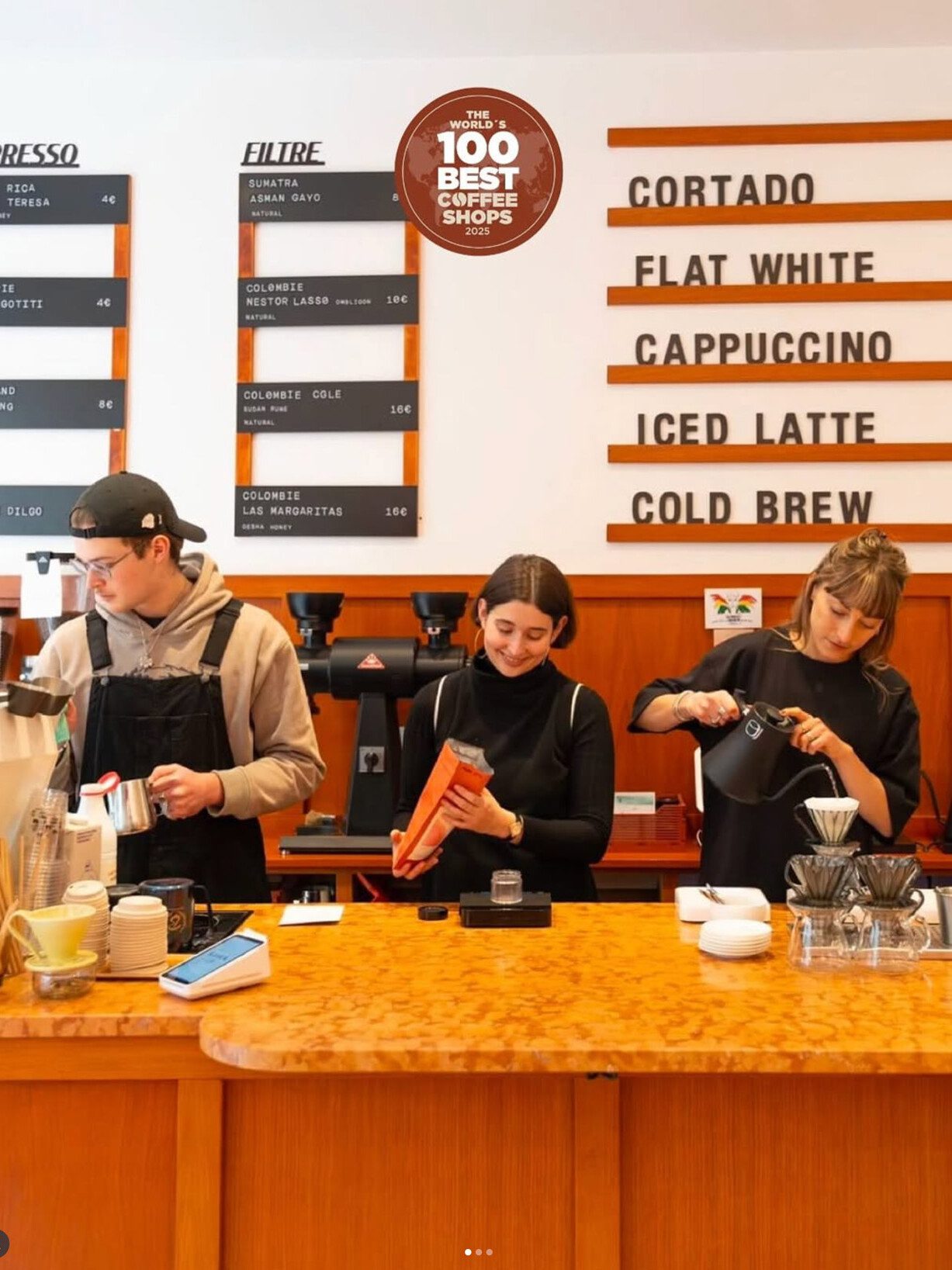 Three baristas prepare drinks behind a coffee bar with a menu board listing espresso and filter options and classic café drinks.