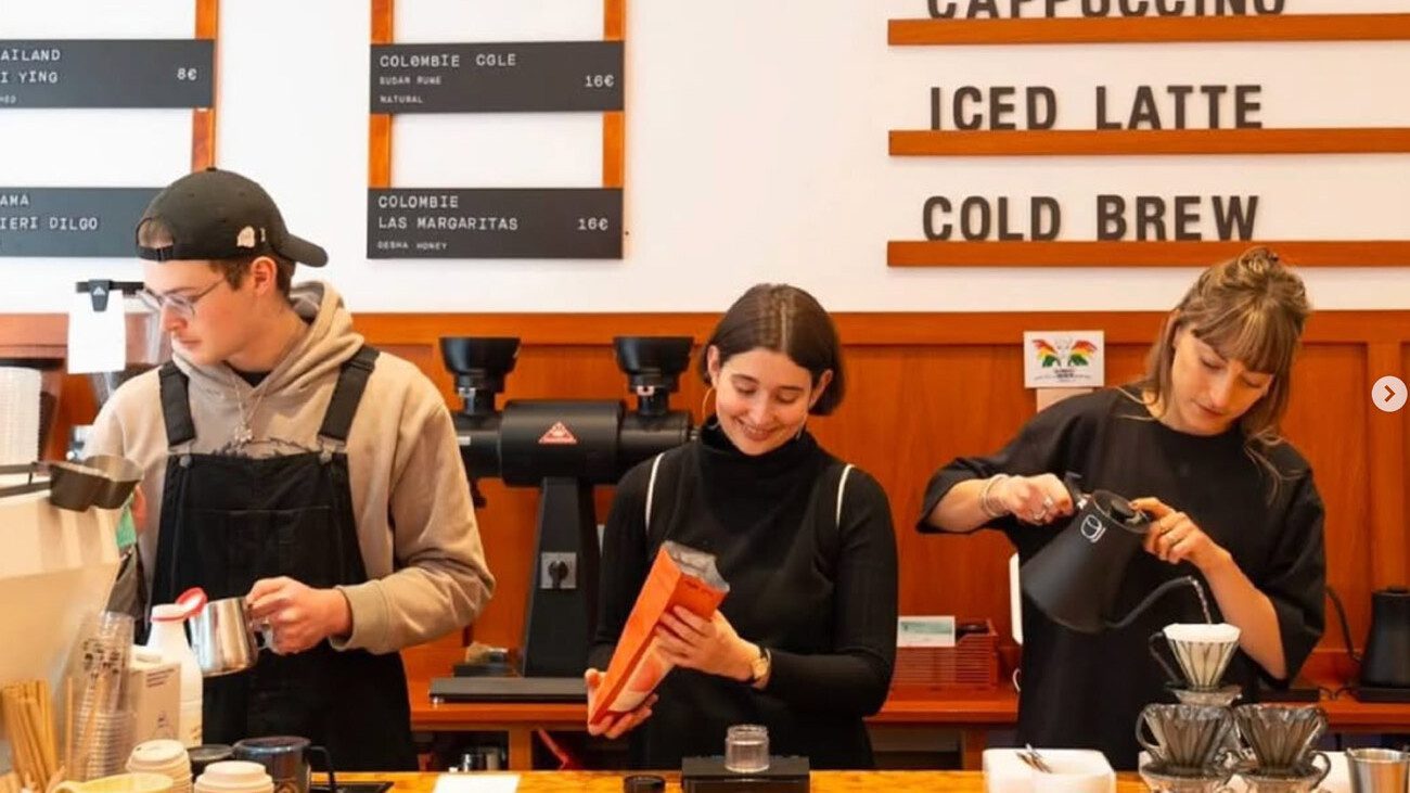 Three baristas prepare drinks behind a coffee bar with a menu board listing espresso and filter options and classic café drinks.