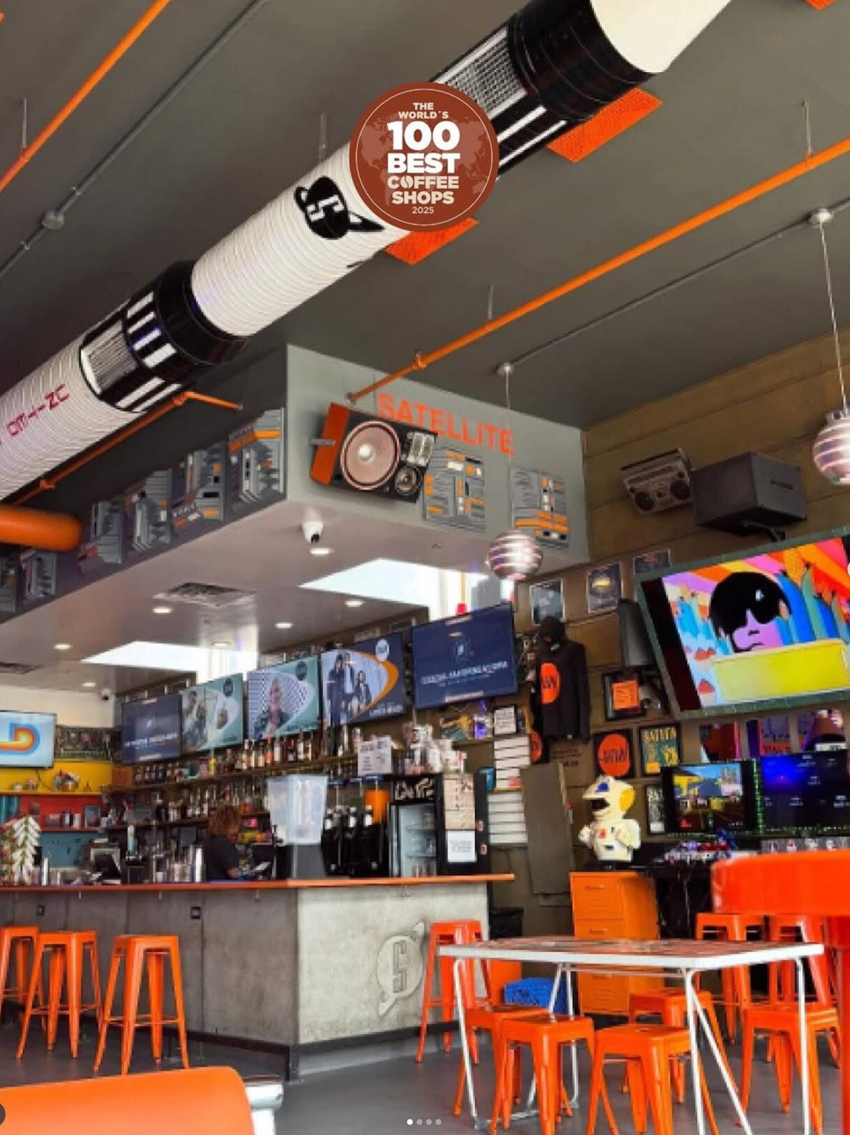 Bright, retro-style coffee shop interior with orange stools and tables, multiple screens behind the counter, and a “Satellite” sign hanging from the ceiling.