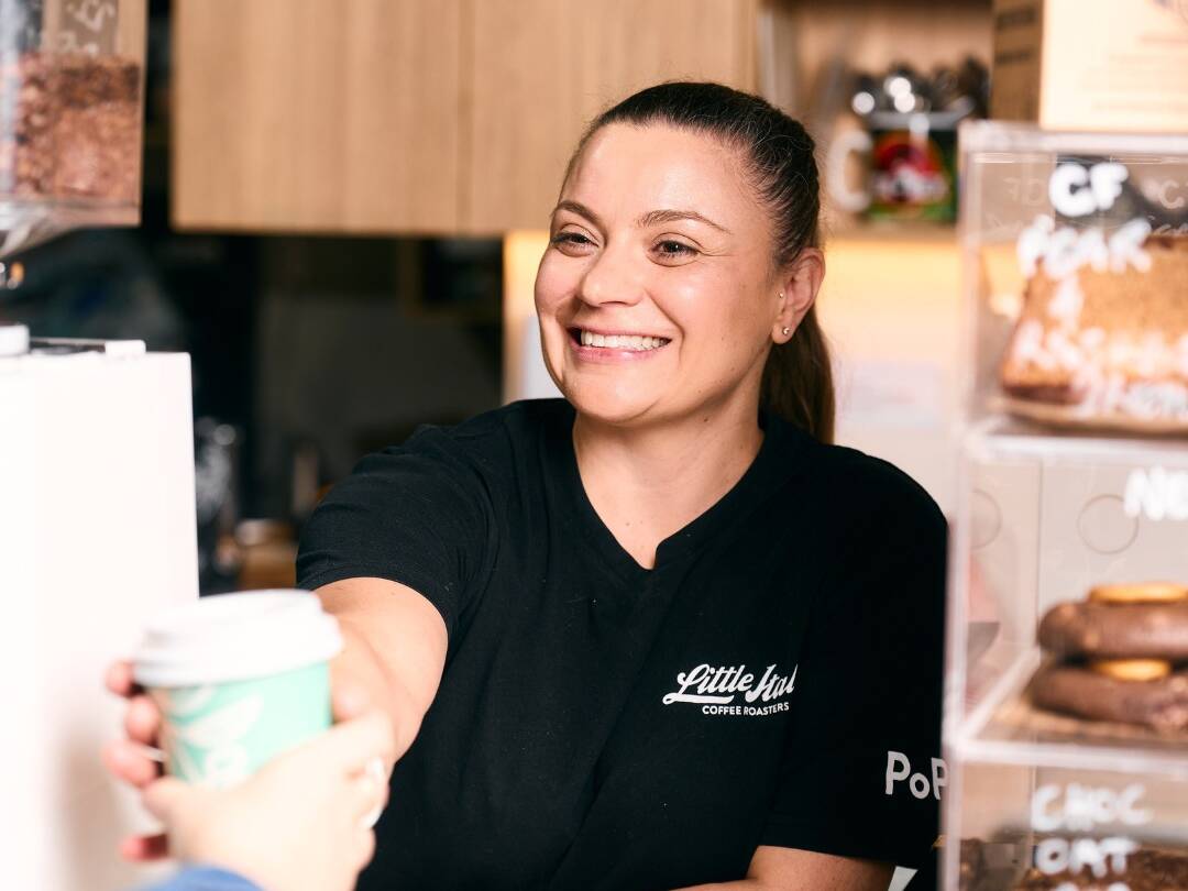 Smiling barista hands a takeaway coffee cup to a customer at a café counter, with pastries displayed nearby.