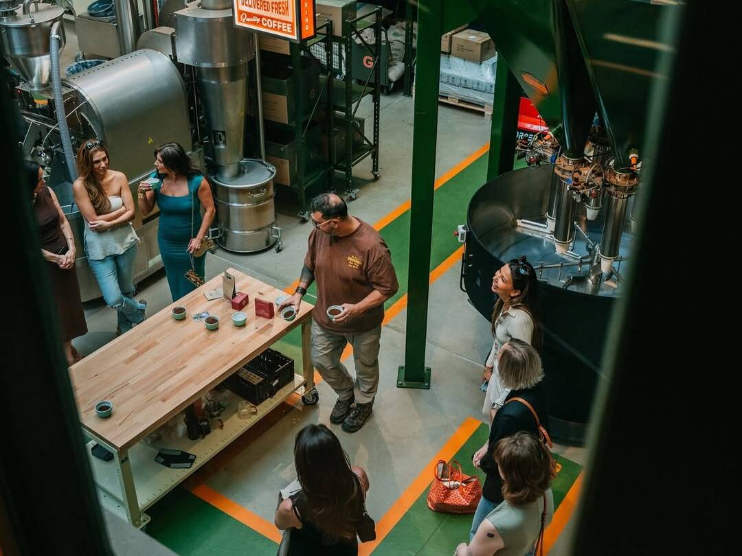 Group of people tour a coffee roasting facility while a guide explains samples at a wooden table surrounded by industrial roasting equipment.