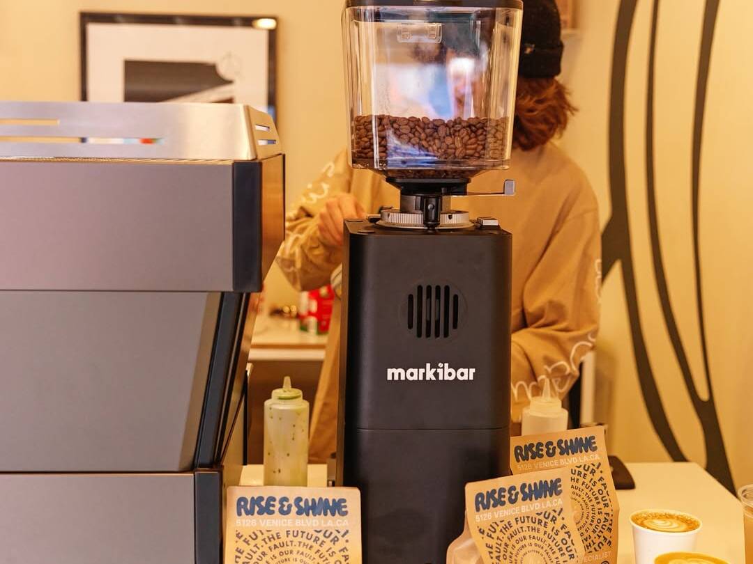 Markibar espresso grinder with coffee beans in the hopper next to an espresso machine, coffee bags, and cappuccino cups on a café counter.
