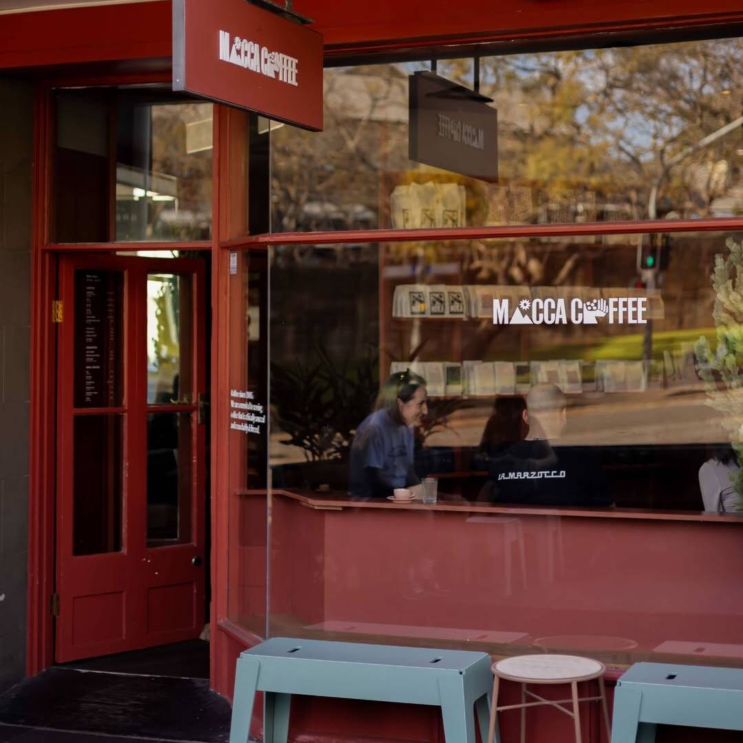 Exterior of a coffee shop with red-framed windows, hanging signage, and customers seated inside by the front window.
