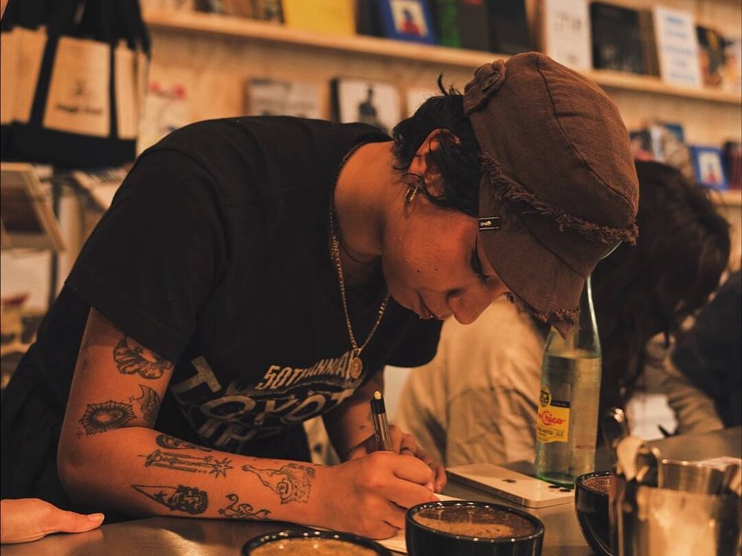 Person wearing a brown cap writes tasting notes during a coffee cupping session, with coffee cups and brewing tools on the counter.