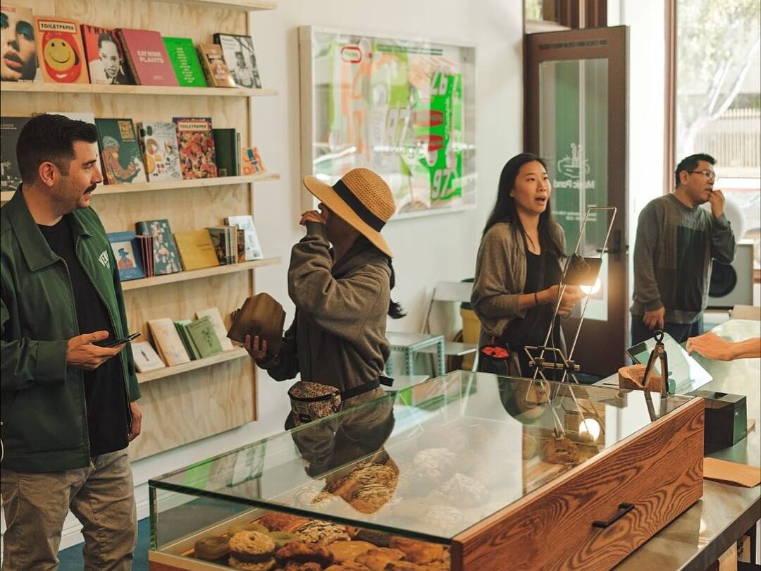 Customers order at a bright café counter with a glass pastry case, books on wall shelves, and baristas serving drinks.
