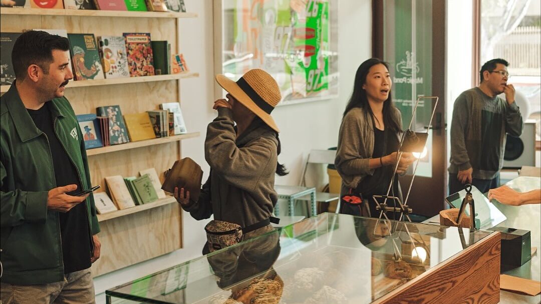 Customers order at a bright café counter with a glass pastry case, books on wall shelves, and baristas serving drinks.