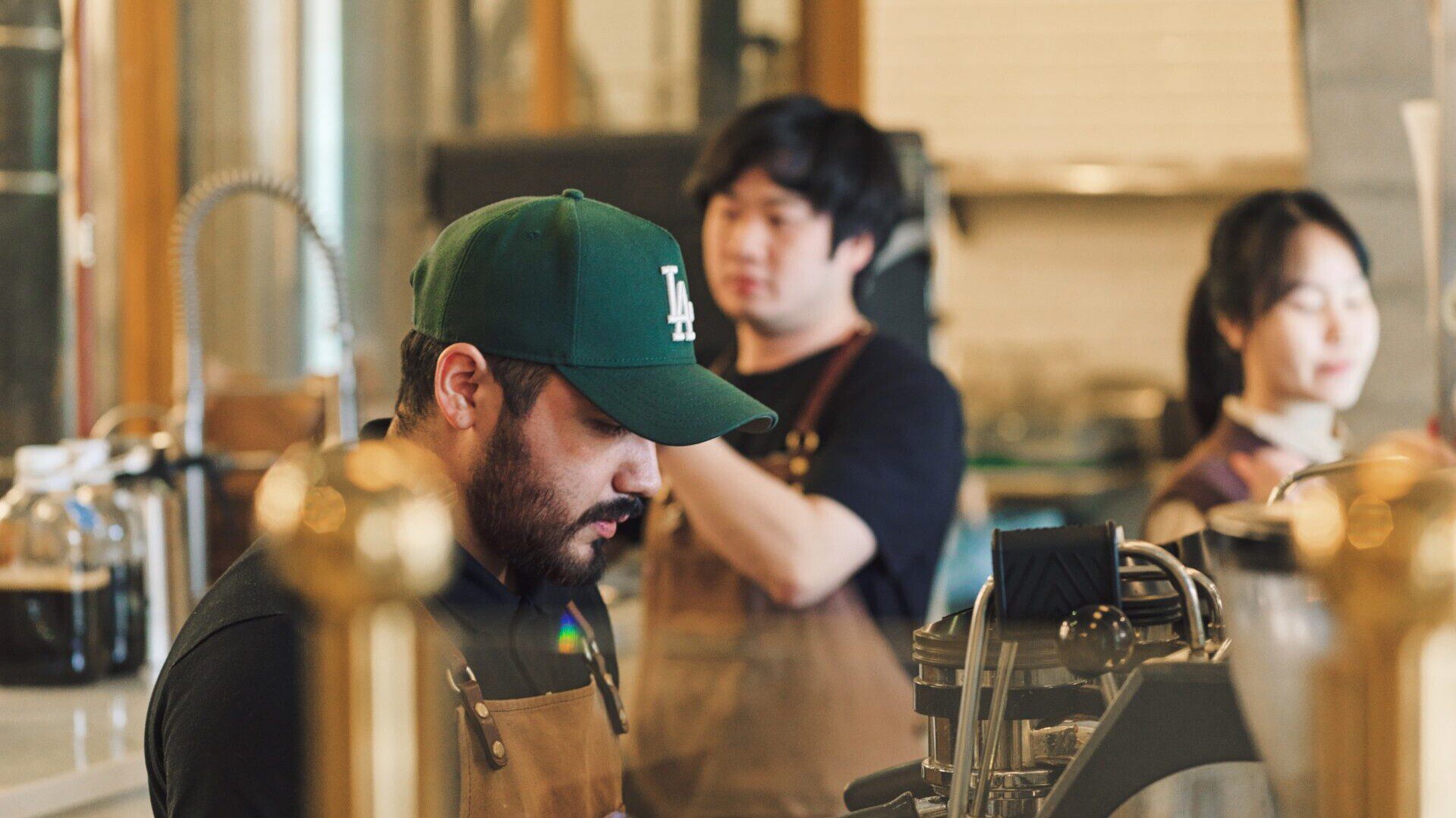 Barista in a green LA cap and brown apron works at an espresso machine in a busy café, with two coworkers blurred in the background.