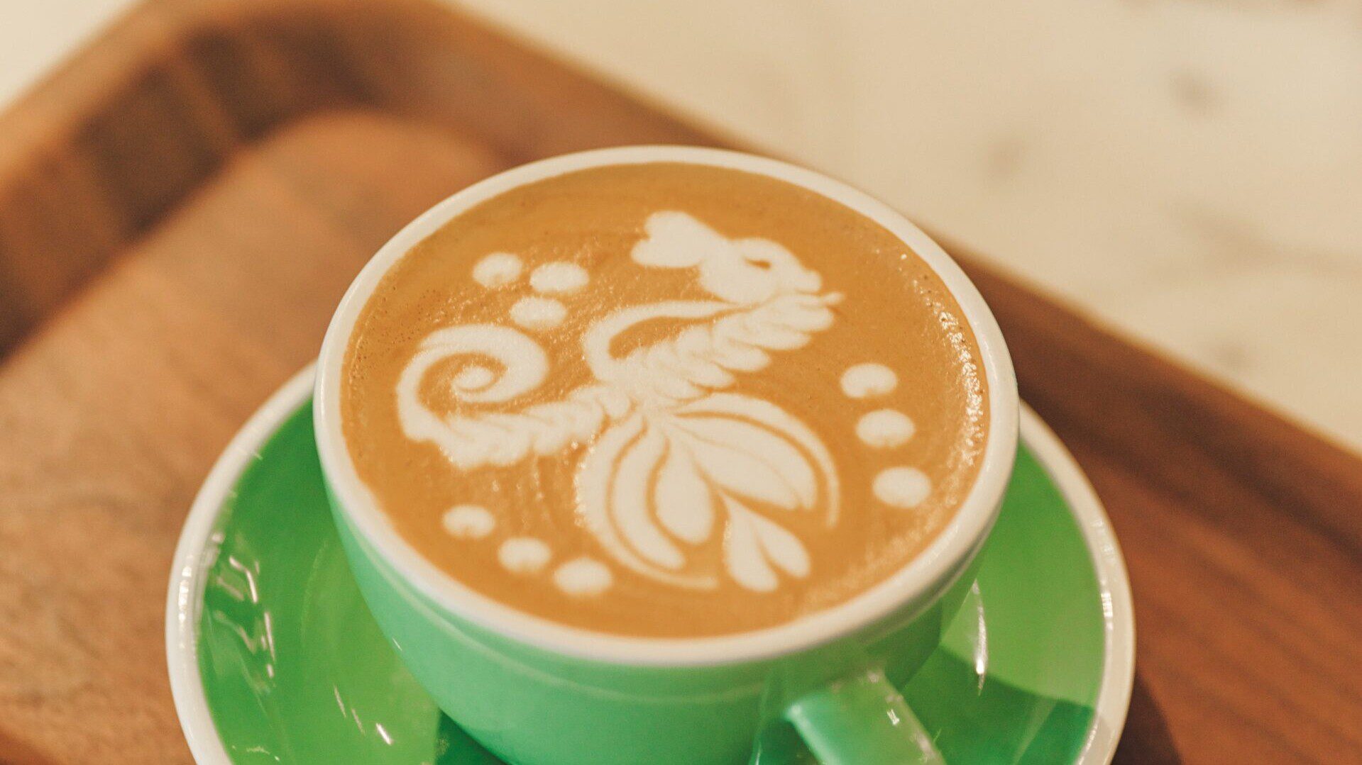 Latte with intricate latte art served in a green cup and saucer on a wooden tray.
